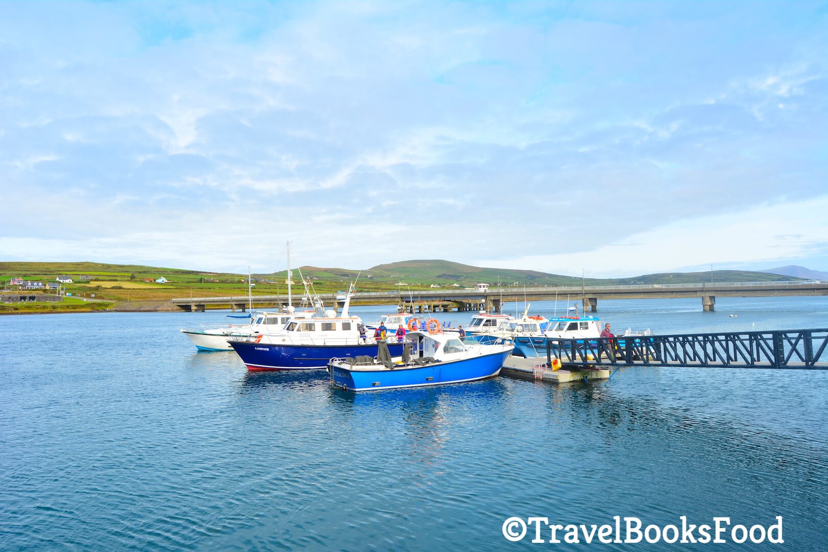 A photo of Portmagee Harbour with crystal clear blue water and few boats with a bridge and some mountains in the distance