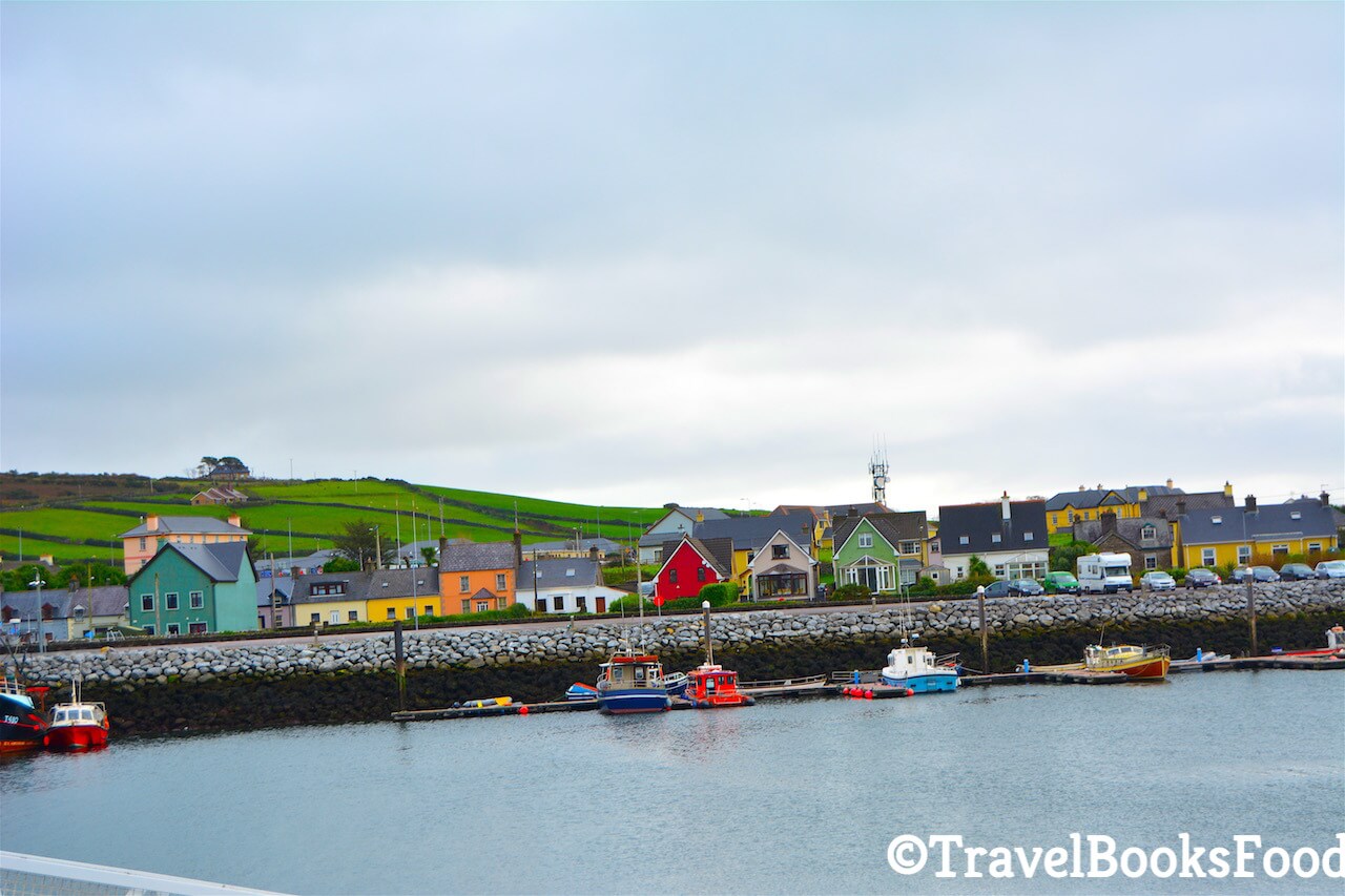 This is a photo of the town of Dingle in Ireland of colourful houses taken from a boat. You can see green fields in the distance