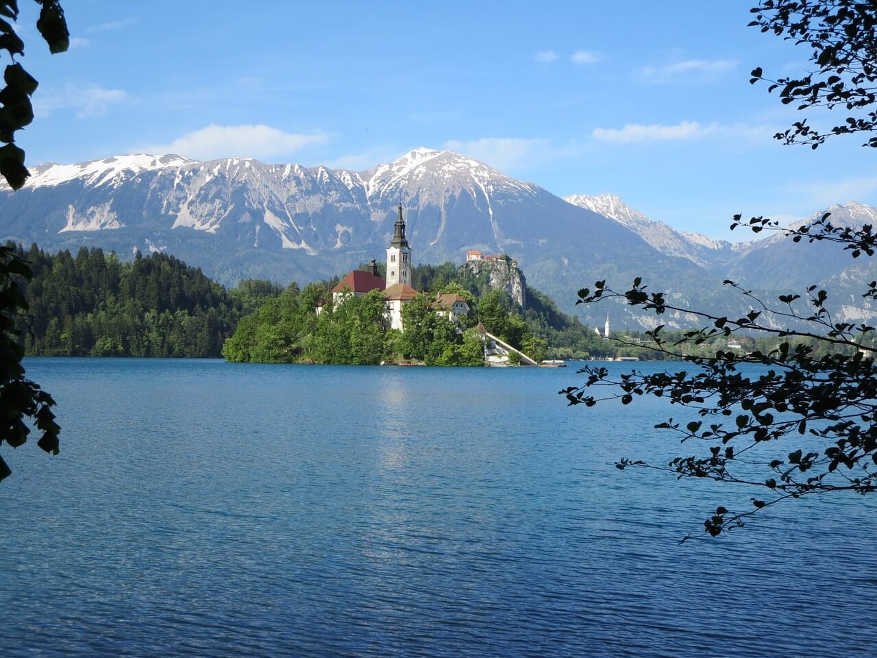 A photo of Lake Bled with a town in the distance and few mountains surrounding it