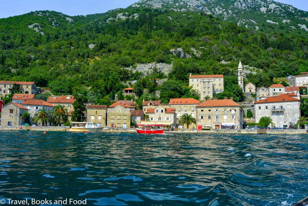 In this photo, you see a coastal town set up against some green mountains with orange roofs and blue water in Perast, Montenegro